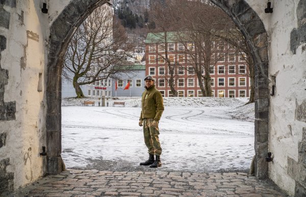 En mann i militærklær står midt i en steinbelagt åpen port. Bak han er det bygninger og trær.