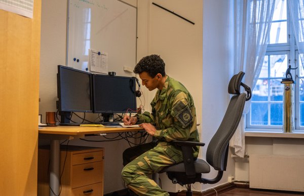 Mann i militærklær sitter ved et skrivebord og skriver i en svart, liten bok.