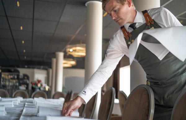 Mann i hvit skjorte og kelnerforkle legger bestikk på et bord.