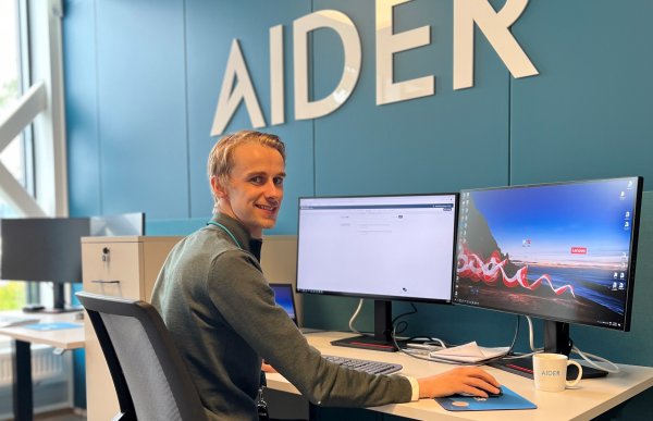 En ung mann sitter på en kontorplass med to store PC-skjermer foran seg på pulten. Han snur seg sidelengs og smiler til kameraet. Han har mørkblondt, kort hår og er ikledd en grønn genser. 
