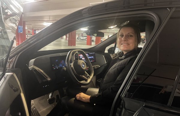 En kvinne kledd i svart er avbildet i førersetet av en svart bil. Hun smiler mot kamera.