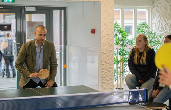 Mann i blå skjorte og grønn dressjakke spiller bordtennis med elever på en skole.