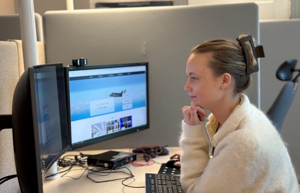 Dame med hvit jakke sitter ved en arbeidspult. Hun ser på to dataskjermer. Damen er på et kontor.