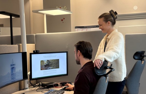 En mann i mørk genser og en kvinne i hvit genser. Mannen sitter på en kontorstol, damen står ved siden av. De ser på to dataskjermer på et skrivebord. De er på et kontor.