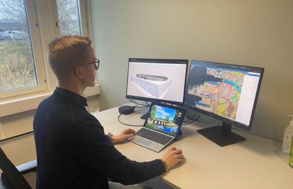 En mørkkledd mann med blondt hår og briller. Han sitter ved et skrivebord. Foran han er en bærbar PC og to store skjermer. På en av skjermene ser vi et kart, og på den andre skjermen ser vi en modell av en bygning.