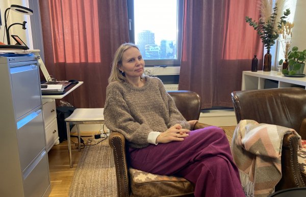 En kvinne med halvlangt blondt hår iført brun strikket genser og rosa bukse sitter i en brun lenestol. Bak henne henger røde gardiner, og ut av vinduet kan vi se noen blokker.