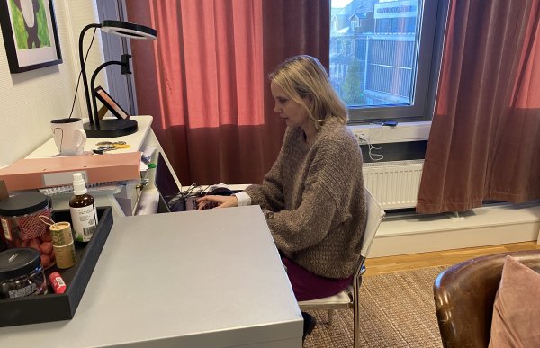 Vi ser en kvinne med halvlangt blondt hår med brun strikket genser og rosa bukse fra siden. Hun sitter ved et skrivebord og skriver på en PC.