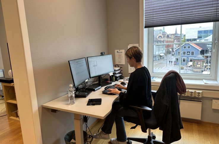 En kvinne sitter ved et skrivebord på kontor og jobber på to dataskjermer. Hun har mørk overdel og lyse jeans, og på stolen henger en jakke. Utenfor vinduet ser man en bygate med bygninger og mennesker.