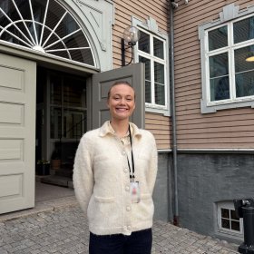Dame med hvit skjorte står foran en beige bygning. Hun har adgangskort rundt halsen. Hun smiler og ser rett i kameraet.