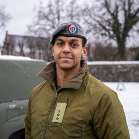 Mann ikledd militære klær og beret og smiler. Bak han skimter man en militærbil.