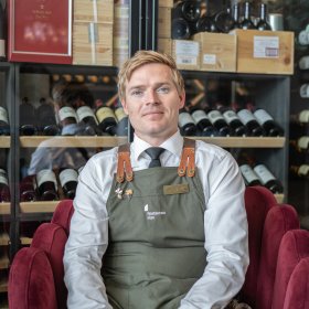 Mann i hvit skjorte og med et grønt kelnerforkle sitter foran et vinskap.