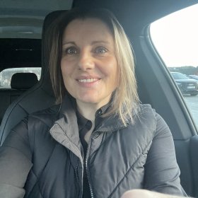 En kvinne med lyst hår smiler mot kamera. Hun sitter inne i en bil. Hun har på seg en svart jakke. Ut av bilvinduet kan man se flere biler.