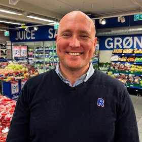 En mann smiler i arbeidsuniform inne i en Rema 1000- butikk