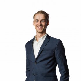En ung mann står rett opp og ned med hendene i lommene på buksen, og smiler til kameraet. Han har mørkblondt, kort hår. Mannen har på hvit skjorte, mørkeblå dressjakke og beige bukser. 