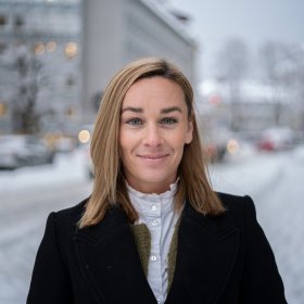 Kvinne med blondt hår i svart kåpe står ute i en gate med snø.