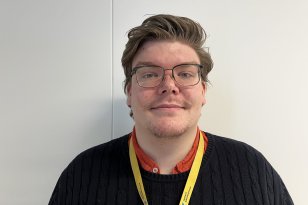 En mann med briller. Han har på svart genser, og har en gul nøkkelholder rundt halsen. Det er en hvit vegg i bakgrunnen.