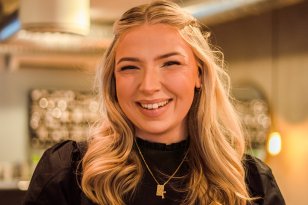 Portrettbilde av frisøren. Hun smiler i kamera, har blondt bølgete hår og sort genser på seg. 