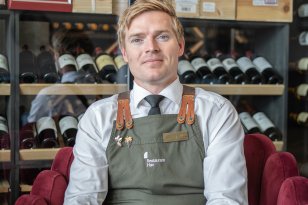Mann i hvit skjorte og med et grønt kelnerforkle sitter foran et vinskap.