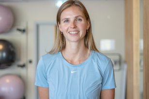 Profilbilde av kvinne med blondt hår og lyseblå trenings t-skjorte som står i et treningsrom med yogaballer.