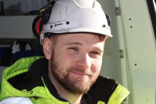 En mann med skjegg smiler til kamera. Han har på seg en hvit hjem og signalgul jakke. 