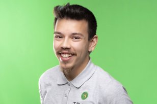 En ung mann med mørkt, kort hår står og smiler stort og vennlig til kameraet. Det er helt grønn bakgrunn. Han har på seg en hvit t-skjorte med krage. Mannen har litt bart og noe skjegg. 