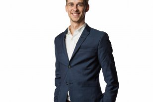 En ung mann står rett opp og ned med hendene i lommene på buksen, og smiler til kameraet. Han har mørkblondt, kort hår. Mannen har på hvit skjorte, mørkeblå dressjakke og beige bukser. 