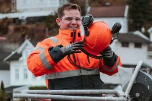 En mann med oransje arbeidsklær står og smiler til kamera. Han holder en oransje vernehjem i hendene. I bakgrunnen sees noen hvite hus. 