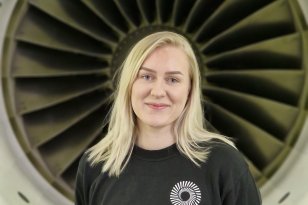 Dame med lyst hår og svart genser. I bakgrunnen er en stor flymotor.
