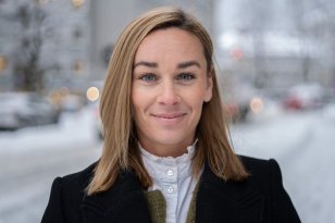 Kvinne med blondt hår i svart kåpe står ute i en gate med snø.