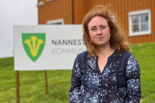 En kvinne står i en gressbakke foran et oransje hus. Hun har gyllent, langt, bølgete hår, briller, og er ikledd en olabukse og en mønstrete mørkeblå bluse. Hun smiler mot kamera.