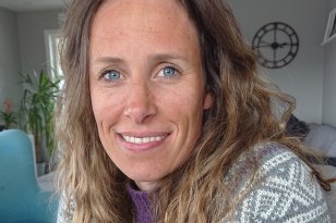 Portrettfoto av dame med brunt, litt krøllete hår. Hun har blå øyne og smiler mot kamera. Hun er ikledd en ullgenser med mariusmønster. Bak henne står det en blå stol.