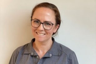 En kvinne ikledd en grå t-skjorte og briller smiler mot kamera. Hun har brunt, langt hår i en hestehale. Bakgrunnen er en hvit vegg.