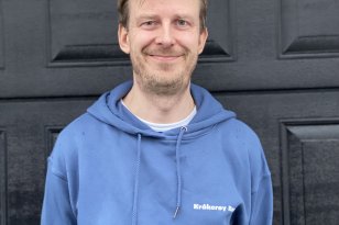 Mann med blå hettegenser står foran en svart dør og smiler.