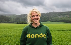 En dame står i en åker og har på seg en grønn genser der det står 'bonde' på. Det er skog i bakgrunnen og himmelen er grå. Damen har blondt går og smiler til kamera
