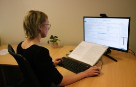 En dame sitter foran en PC, foran skjermen ligger en åpen bok med tekst 