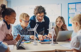 En lærer forklarer noe for en gruppe på fire elever i barneskolealder. Midt på bordet ligger en iPad, som læreren peker mot.