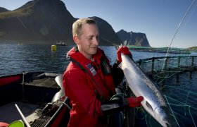 Fiskeoppdretter som holder opp en fisk han har henta opp fra merden bak.