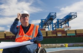 Mannlig skipsmegler i rullestol studerer logistikkplaner på kaia ved et fraktskip.