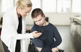 Kvinnelig vernepleier hjelper mann i rullestol med en mobiltelefon.