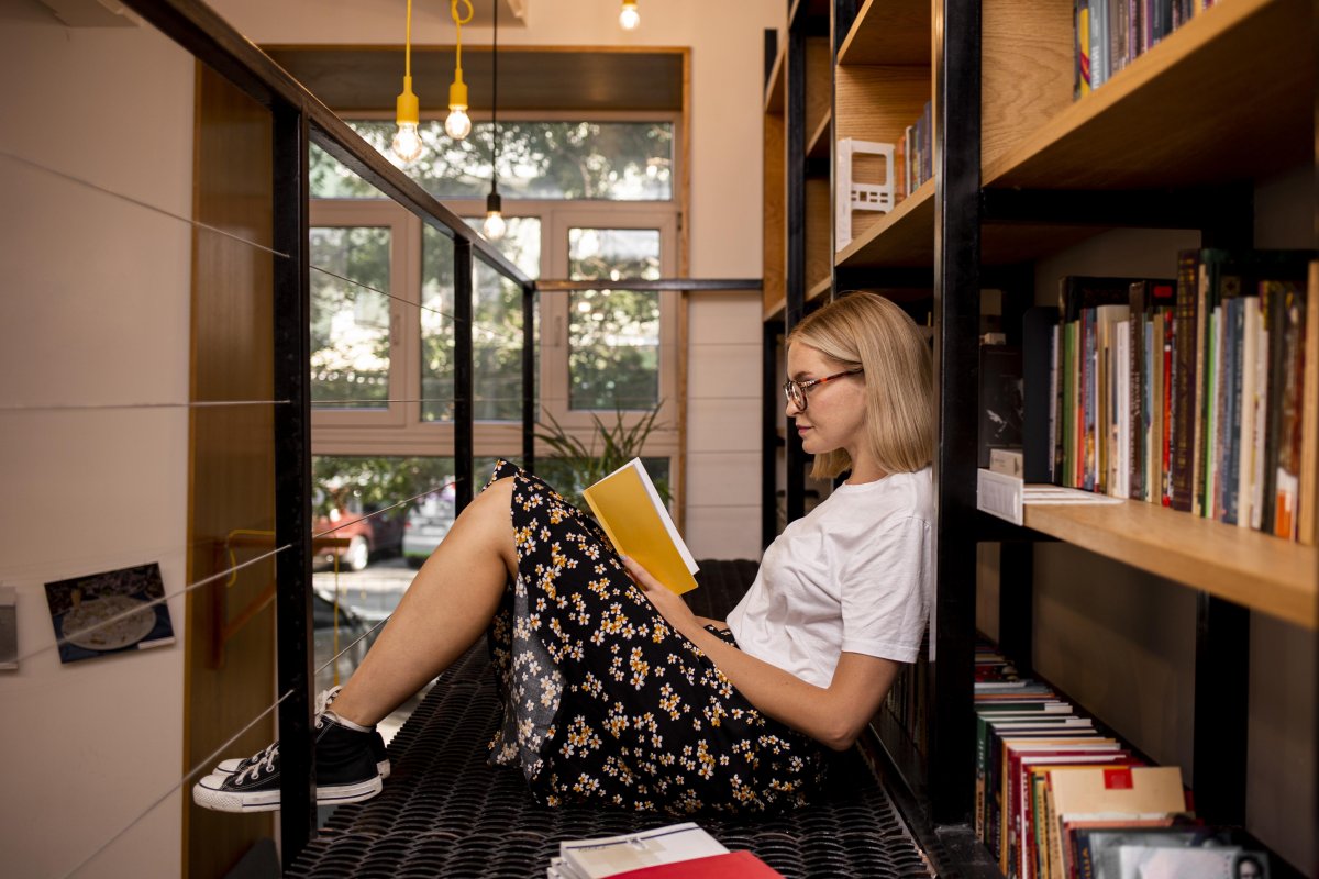 En ung kvinne sitter i et bibliotek og leser en i en bok. Hun er kledd i skjørt og hvit t-skjorte og bruker briller.