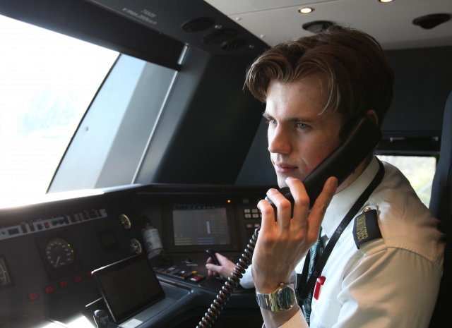 Ung mann i uniform snakker i telefonen. Han sitter i førerrommet på et tog.