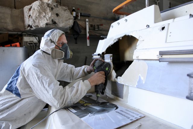 Komposittbåtbyggar Jack André Jakobsen ikledd verneklede og maske, med pussemaskin i hendene. Står bøygd over ein båt med eit hol. 