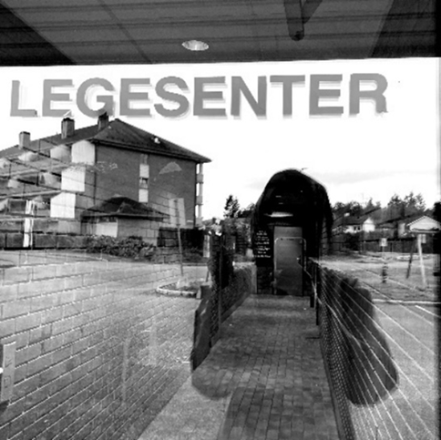 Bilde av et vindu med skriften "Legesenter", i gjennskinnet ser man den kvinnelige fotografen.
