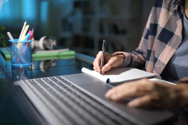 Bilde av hender som skriver på en blokk med en PC ved siden av